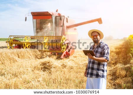 Picture of a smiling bearded farmer with a tablet in a wheat field in front of a working combine harvester looking into the camera.