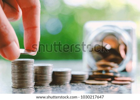 Hand saving a coin on the stack of money on blurred the glass jar and green natural background for investment, business and finance concept