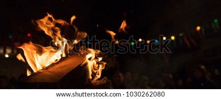 Bonfire with colorful flames and bokeh lights. Big blaze with firewoods. Blurred people on night background. Close up view with details, banner, space for text.