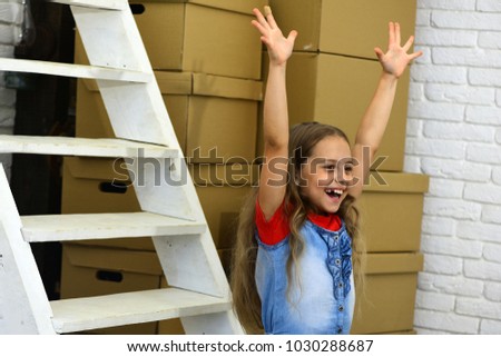 Kid stands by pile of cardboard boxes and white ladder. Girl with fair wavy hair on room background. Child with excited face puts hand up in joy. New home and moving in concept