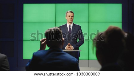 View from behind a production team in a studio sitting listening to a presenter talking in front of a green screen then breaking into applause clapping.