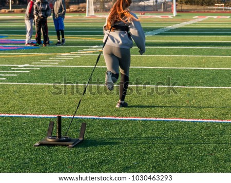 A high school athlete is running away from the camera pulling a weighted sled for strength and speed work at practice.