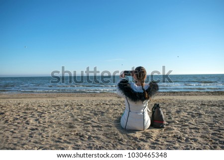 back view of a girl in a white warm jacket sitting on the sand and taking pictures of the sea with a mobile phone. View of the model against the blue sea and sky