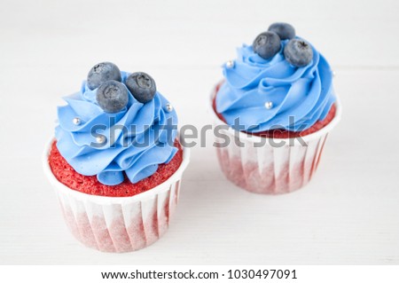 Cupcake red velvet with blue whipped cream decorated with blueberry, silver confectionery balls on white wood table. Picture for a menu or a confectionery catalog.