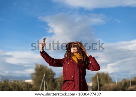 young beautiful happy woman taking a selfie with her mobile phone over blue sky background in the city. Autumn or winter season. Lifestyle
