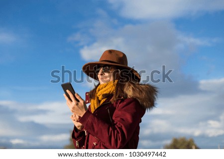 young beautiful happy woman using mobile phone. She is wearing winter clothes. Blue sky background . Autumn or winter season. Lifestyle
