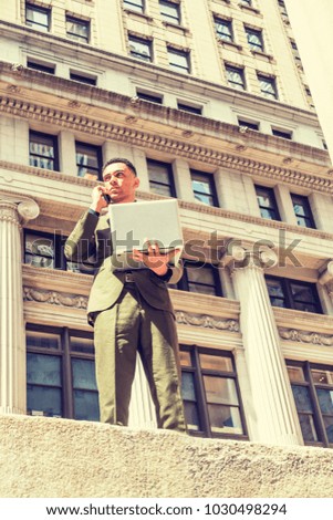 American Businessman traveling, working in New York, wearing green suit, standing on street with vintage buildings, working on laptop computer, calling on cell phone. Filtered effect.
