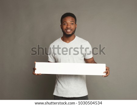 Picture of young smiling african-american man holding white blank board for advertisement on grey background, copy space
