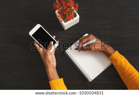 Black female hands making notes in notebook and holding smartphone. Top view of african-american hands, laptop keyboard on a wooden table background. Education, business, technology concept