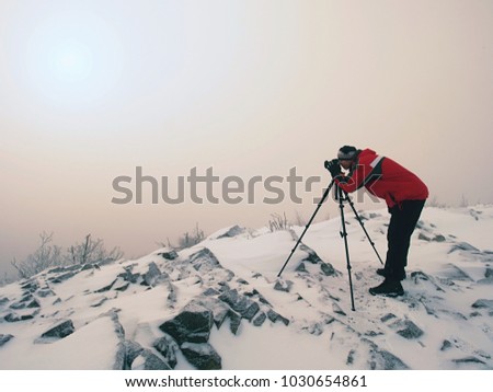 Photographer takes picture of freeze autumnal daybreak, rocks covered with fresh powder snow. Stony rock peak increased from thick mist. 