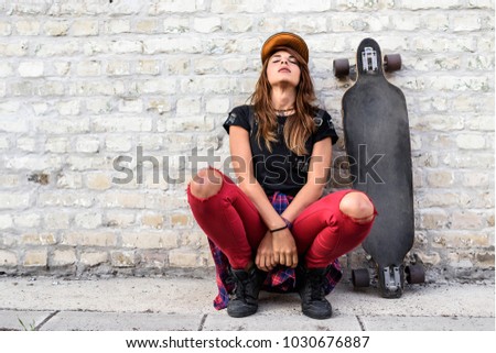 Cute urban girl with longboard sitting sitting next to a brick wall outdoors