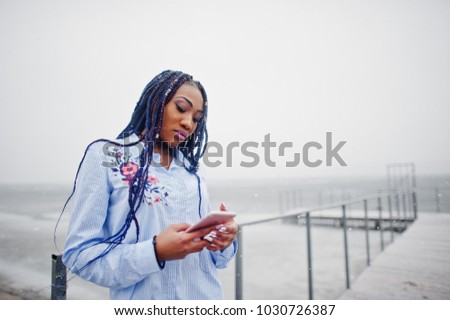 Stylish african american girl with dreads holding mobile phone at hand, outdoor on pier against frozen lake at snowy weather.