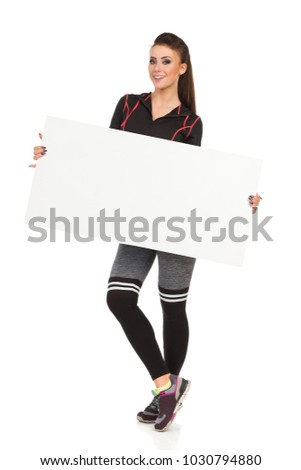 Beautiful young woman in fitness clothes is standing, holding white poster, looking at camera and smiling. Full length studio shot isolated on white.