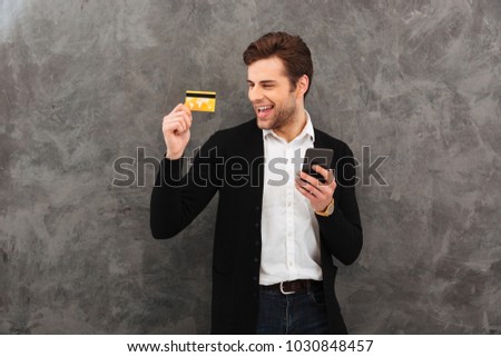 Picture of young businessman standing over grey wall background. Looking aside chatting by mobile phone holding credit card.