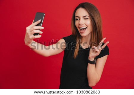 Cool classy woman in black t-shirt partying and taking selfie with victory sign on her cell phone isolated over red background