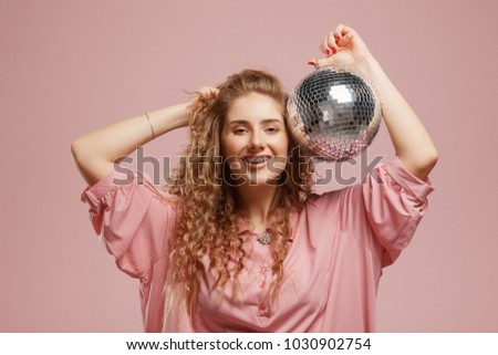 Closeup of beautiful smiling young woman with sparkles in hair and on face posing near disco ball over pink background. Concept party, holiday, surprise