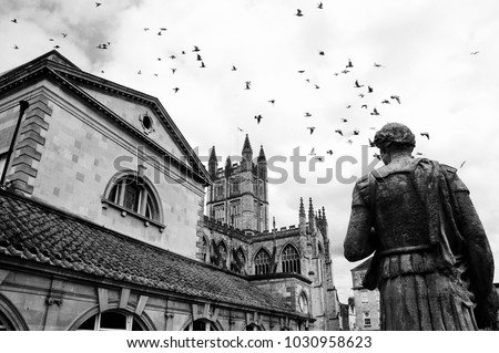 Bath (England, UK) Stone statue of the roman in Antique Roman Baths complex, flying birds in sky and Abbey Cathedral at background. City of Bath is a UNESCO World Heritage Site. Black and white photo.