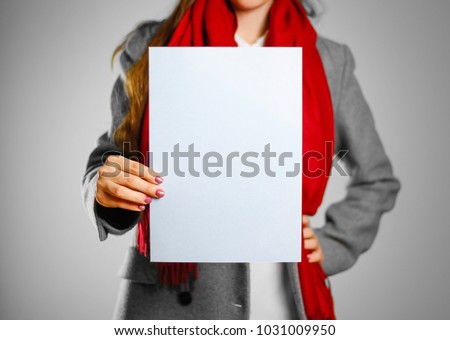 A girl in grey coat and red scarf keeps a light blue clean blank sheet of A4. Isolated on grey background.