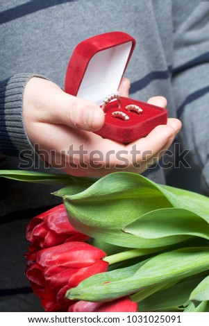 Gifts for loved ones. The men is holding a bouquet of red tulips in her hand. In the other hand, an open velvet box of red color, in which a ring and earrings.