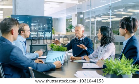 Senior Executive Explains Company's Vision and Potential to His Employees. They are Sitting at Big Table in Meeting Room. TV Screen on the Wall Shows Corporate Growth. Royalty-Free Stock Photo #1031044153