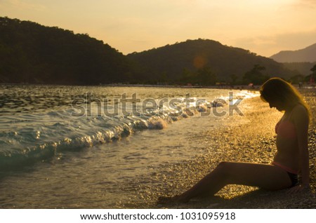 Girl in pink bikini swimwear is sitting on the sea beach. Beautiful sunset at the seaside with mountains on background. A stream of light in evening sky. Gorgeous woman silhouette. Ocean beach.