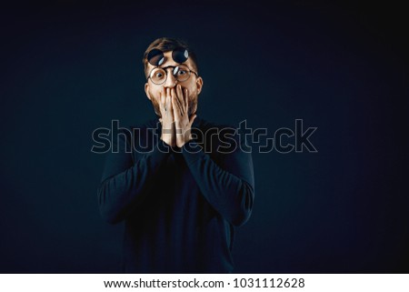Young bearded man in black covering his mouth with hands looking with surprise at camera wearing round flip-up glasses on black. Emotion, silence and people concept.