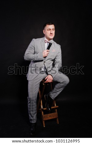 Handsome man in gray suit with microphone against black background on studio sitting on chair. Toastmaster and showman.