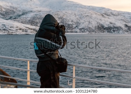 Man photographs seascape, standing on deck of a ship. Sea and snow-capped mountains in the background.
