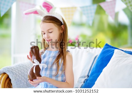 Cute little girl wearing bunny ears eating chocolate Easter rabbit. Kid playing egg hunt on Easter. Adorable child celebrate Easter at home.