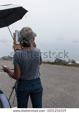 A female artist is standing in a parking lot with an unbrella protecting her work, painting the foggy water scene in front of her.