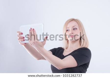 Woman making selfie with tablet on white isolated background. Close up of young blonde beautiful girl in black dress looking in white tablet in her hands

