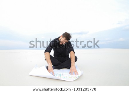 Student of history department talk on smartphone, guy doing map for graduate work in secluded place asking groupmate for advice. Young fair-haired bearded man sitting on sand unwrapping whatman pape