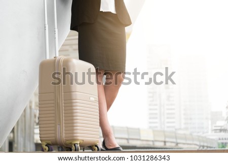 Low body section portrait of a woman with a luggage standing at a transportation terminal ready to travel. Image with blurred city view in the background.