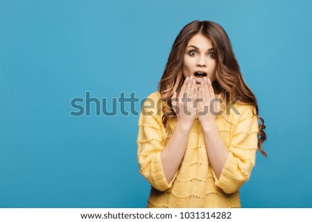 Portrait of young lady in yellow sweater with shocked facial expression on blue background