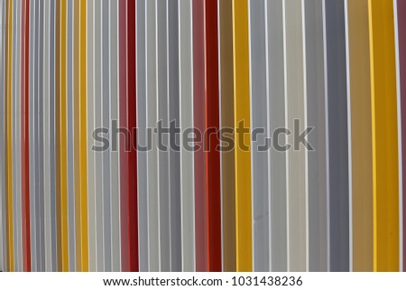 Close up outdoor view of a colorful modern gate made of iron vertical bars. Pattern with lines of different colors: yellow, red, white, gray, orange. Geometric image with striped elements and shadows.