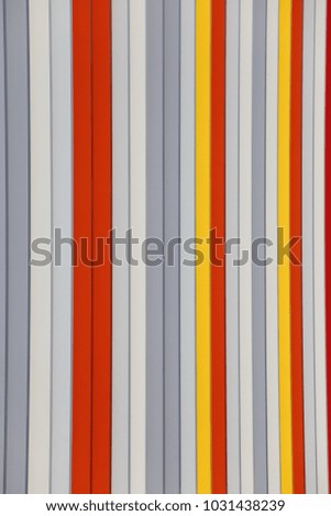 Close up outdoor view of a colorful modern gate made of iron vertical bars. Pattern with lines of different colors: yellow, red, white, gray, orange. Geometric image with striped elements and shadows.