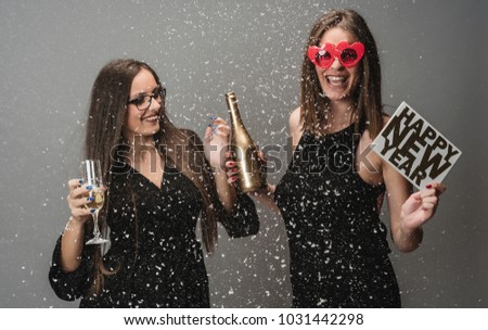 Two female friends celebrate NEW YEAR with confetti and balloons holding glass and bottle of champagne. isolated  
