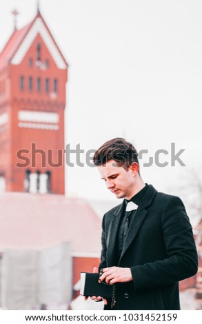 Portrait of handsome young catholic priest reading the prayer book against church background.