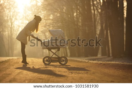 Silhouette of young woman in white coat and black hat with pompom standing elegant in sun on footpath and looking at child in baby stroller on backgroung on sunny winter park, childcare and love