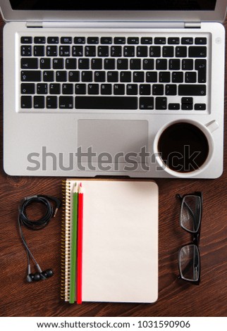 Top view of laptop and notepad with green and red color pencils, black glasses and headphones on rustic brown wood desktop and cup of hot coffee.