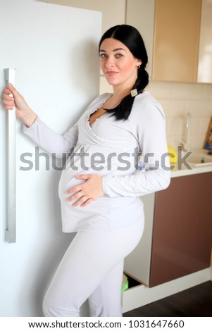The beautiful pregnant woman costs near the refrigerator.