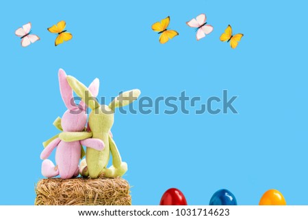 Two rabbits on a straw bale with easter eggs and butterflies in front of a blue background
