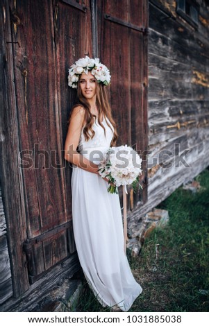 Beautiful girl in long white dress with wreath on the head and bouquet of white peonies in hands, old building in rustic style on background