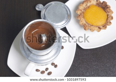 Vietnamese coffee maker is equipped on a cup. It is filled with ground coffee and pour boiling water. There is a cake on the saucer.