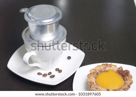 Vietnamese coffee maker is equipped on a cup. It is filled with ground coffee and pour boiling water. There is a cake on the saucer.