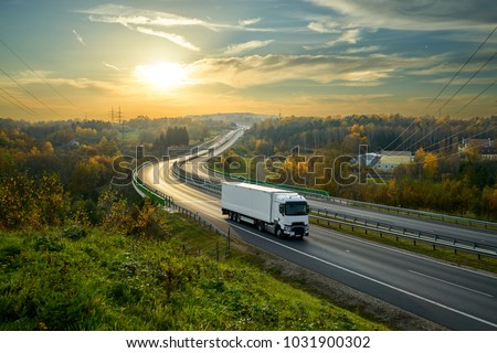 White truck driving on the highway winding through forested landscape in autumn colors at sunset Royalty-Free Stock Photo #1031900302