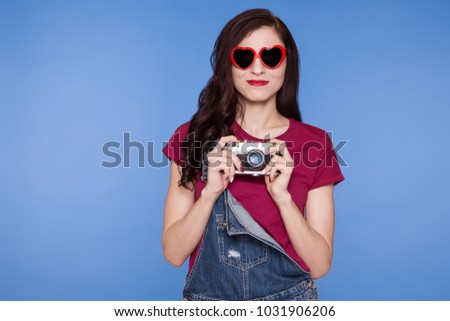 Beautiful brunette woman holding a camera, SLR and smiling. On the face is a sunglasses heart. Retro image. Blue background.
