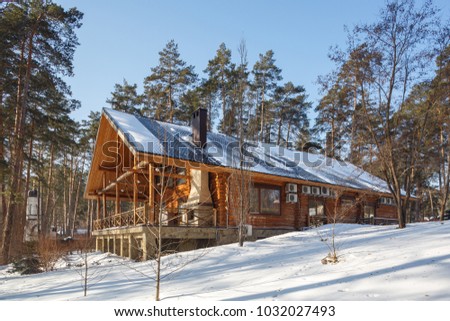 snow covered traditional wooden cottage in chalet style on the hill around pine trees, snow covered, blue sky, sunny day big wooden terrace with fire place with big chimney