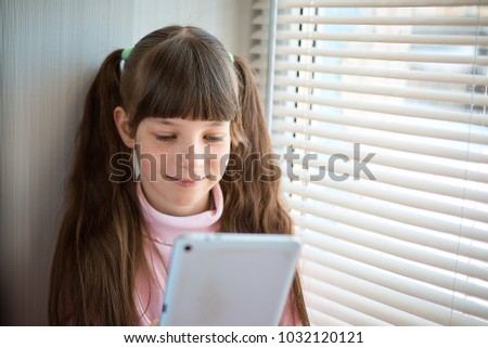a little girl with freckles and blue eyes sitting by the window and using a tablet