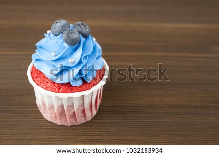 Cupcake red velvet with blue whipped cream decorated with blueberry, silver confectionery balls on dark wood table. Picture for a menu or a confectionery catalog.